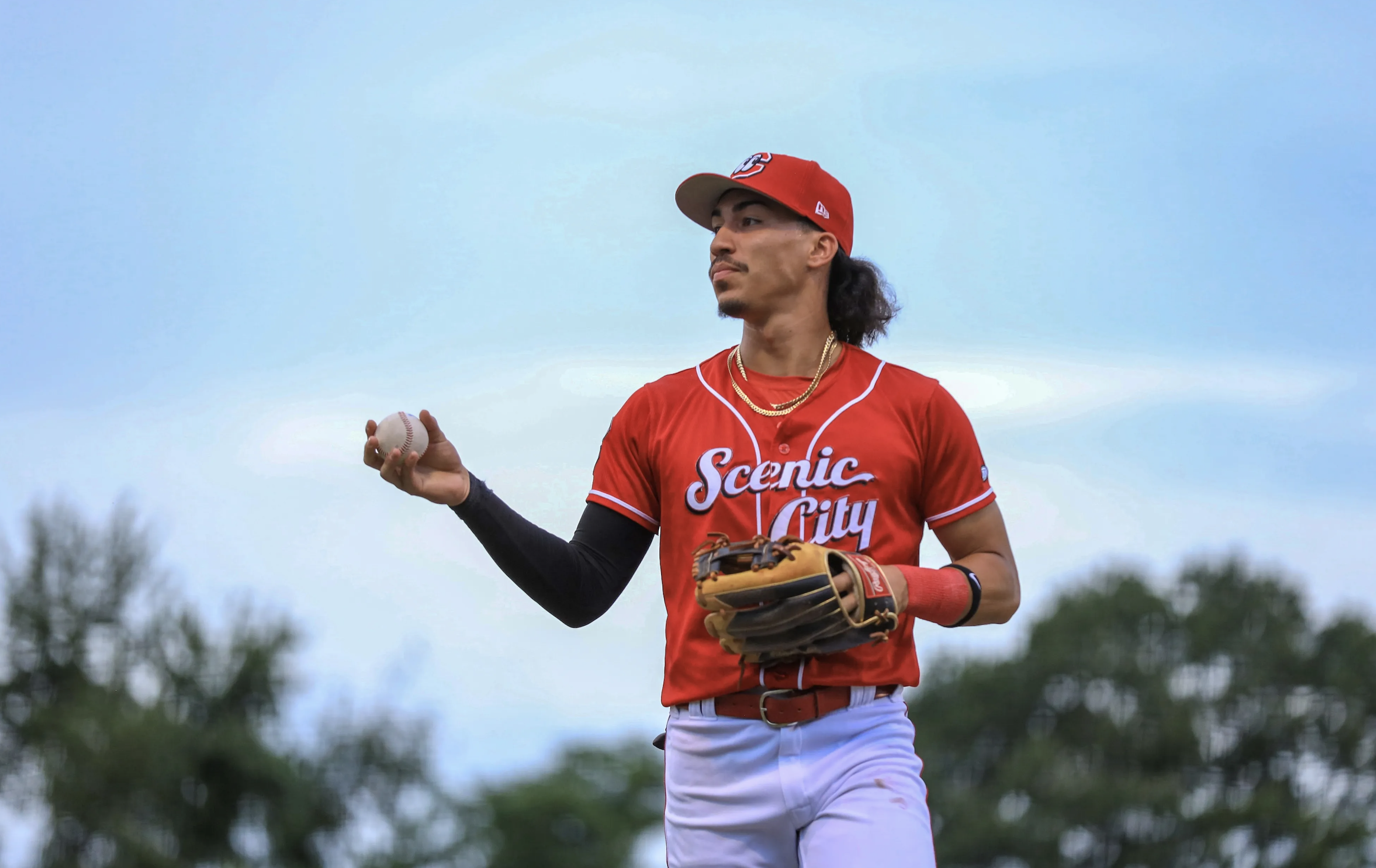 Edwin Arroyo, in a red Scenic City (Chattanooga Lookouts) jersey and cap, holds a baseball in his right hand and a glove in his left, looking off into the distance against a soft blue sky.